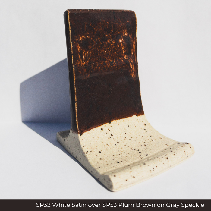 SP32 White Satin glaze over SP53 Plum Brown glaze on a Gray Speckle clay test tile, dark brown with amber streaks