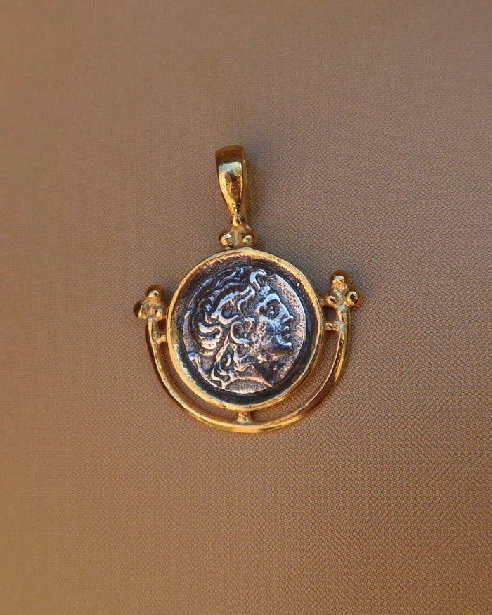 A gold pendant featuring an antique-style circular coin medallion with a raised classical bust of Alexander the Great, displayed on a textured brown background. 18k gold-plated sterling silver 925 made in Greece, Greek coin jewelry