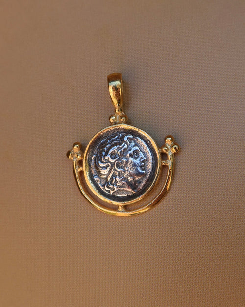 A gold pendant featuring an antique-style circular coin medallion with a raised classical bust of Alexander the Great, displayed on a textured brown background. 18k gold-plated sterling silver 925 made in Greece, Greek coin jewelry
