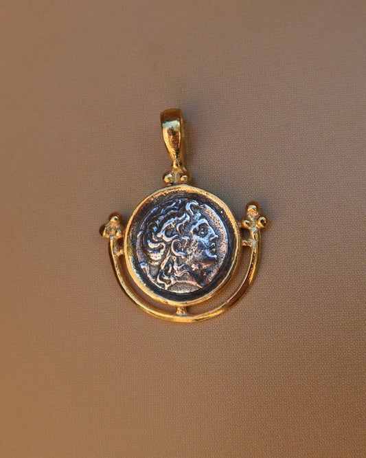 A gold pendant featuring an antique-style circular coin medallion with a raised classical bust of Alexander the Great, displayed on a textured brown background. 18k gold-plated sterling silver 925 made in Greece, Greek coin jewelry