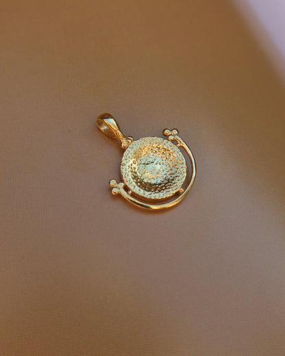 The back of a gold-plated pendant featuring a textured circular center with a curved outer band, displayed on a smooth, warm brown surface.