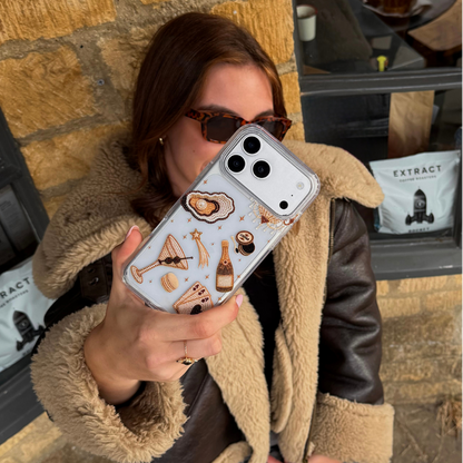 A woman wearing a brown aviator jacket taking a photo with her phone in a cafe. The phone has a clear phone case with illustrations of a gold-toned cocktail, playing cards, and clam with pearl.