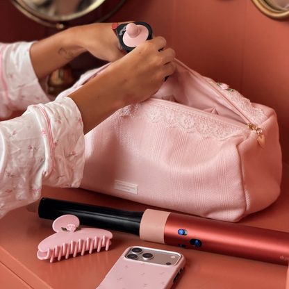 A pink hair tool bag with white lace trim and a gold zipper, resting on a peach-colored cabinet next to a person's hand. A matching pink hair claw clip is in the foreground.