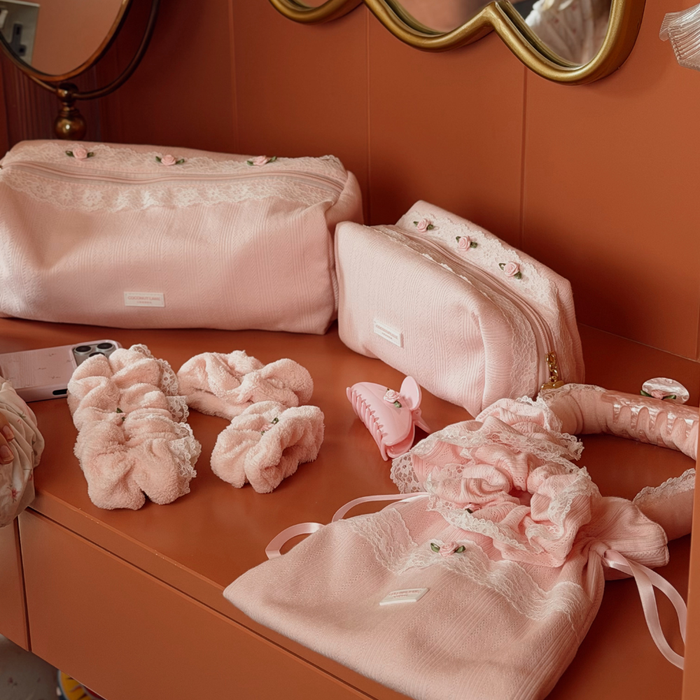 A coordinated set of pink beauty accessories arranged on a warm terracotta vanity surface, including two soft pouches (makeup bag and hair toool bag), pink scrunchies, a hair clip, a ruffled drawstring bag, and a heatless curls set with lace trim and a pink claw clip, with matching pink scrunchies.
