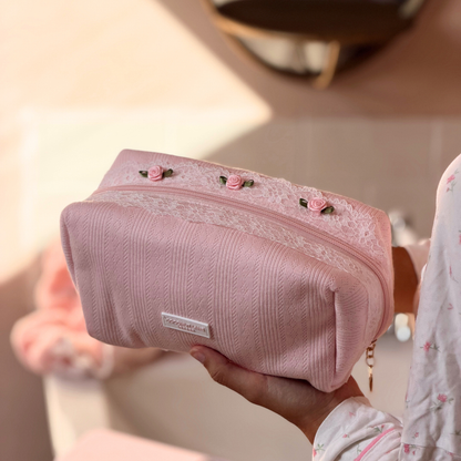 A person holds a light pink cosmetic pouch with a textured fabric body, a white lace trim, and embroidered pink roses on top.