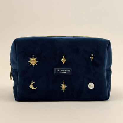 Stardust After Dark makeup bag. Dark navy blue velvet material with gold zip, and sewn-on gold celestial-themed charms including a sun, moon, star, diamond shape and 11:11 charm.