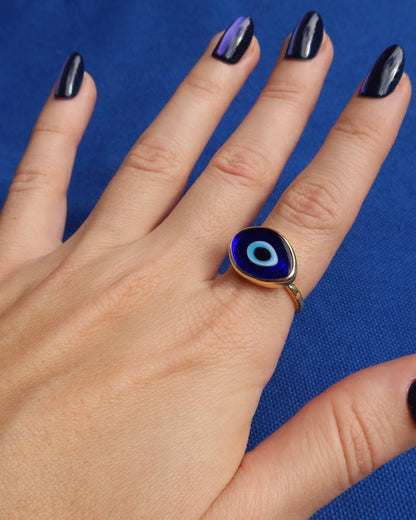 Woman's hand wearing a dark blue Murano glass evil eye ring with a gold band against a blue background, Greek evil eye jewelry