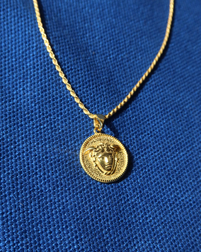 A gold-tone chain necklace with a circular pendant featuring a Medusa-style head motif, displayed on a textured blue fabric background. Greek mythology jewelry