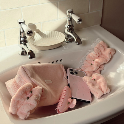 A collection of soft pink bath accessories arranged in a white sink, including plush bath towels, a claw hair clip, a phone case, and a scrunchie, with chrome faucets and a tiled wall in the background.