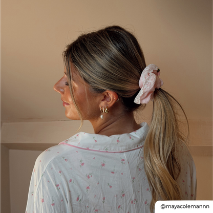 A woman shown from the back and side profile wearing a light pink floral scrunchie in her ponytail, paired with a white floral pajama top. The background is a solid warm beige wall.