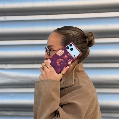 A woman with glasses and her hair in a bun holds a phone with a decorative  autumn-themed case featuring illustrations of pumpkins, a crescent moon, and a slice of pumpkin pie.