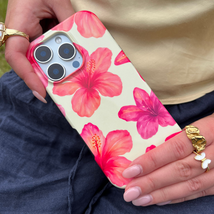 Hand holding a phone with a yellow hibiscus floral Coconut Lane tough case; gold rings and manicured nails visible.