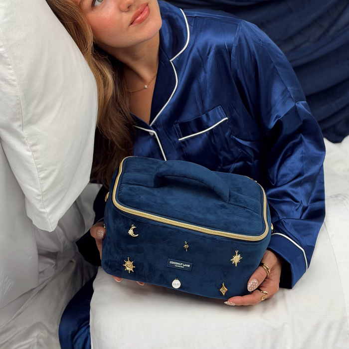 Woman wearing satin navy blue pyjamas. She is holding a stardust After Dark Vanity Case. A dark blue navy velvet vanity case with gold zip, and sewn-on gold celestial-themed charms. Including a moon, sun, star, diamond shape and 11:11 charm.